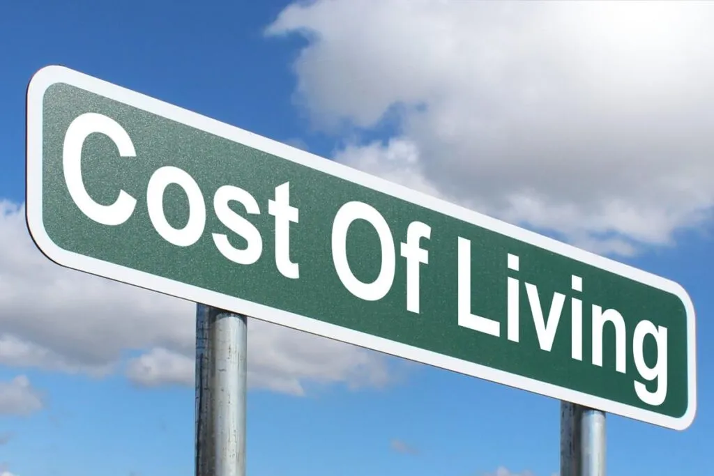 Current image: A green street sign with white borders displaying the text "Cost Of Living", mounted on poles against a blue sky with clouds background.